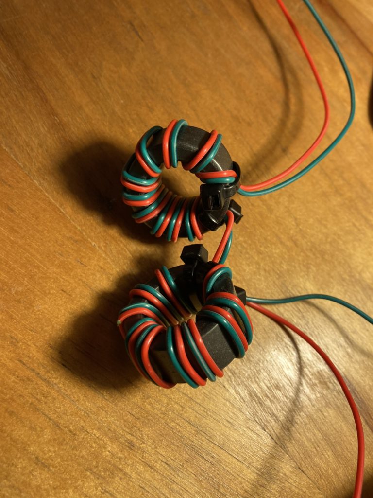 Two 1.25" toroids sitting on a butcher block maple surface, illuminated by warm lamplight. The toroids are wound in series with 26 AWG PVC coated wire, alternating green and red in bifilar windins.