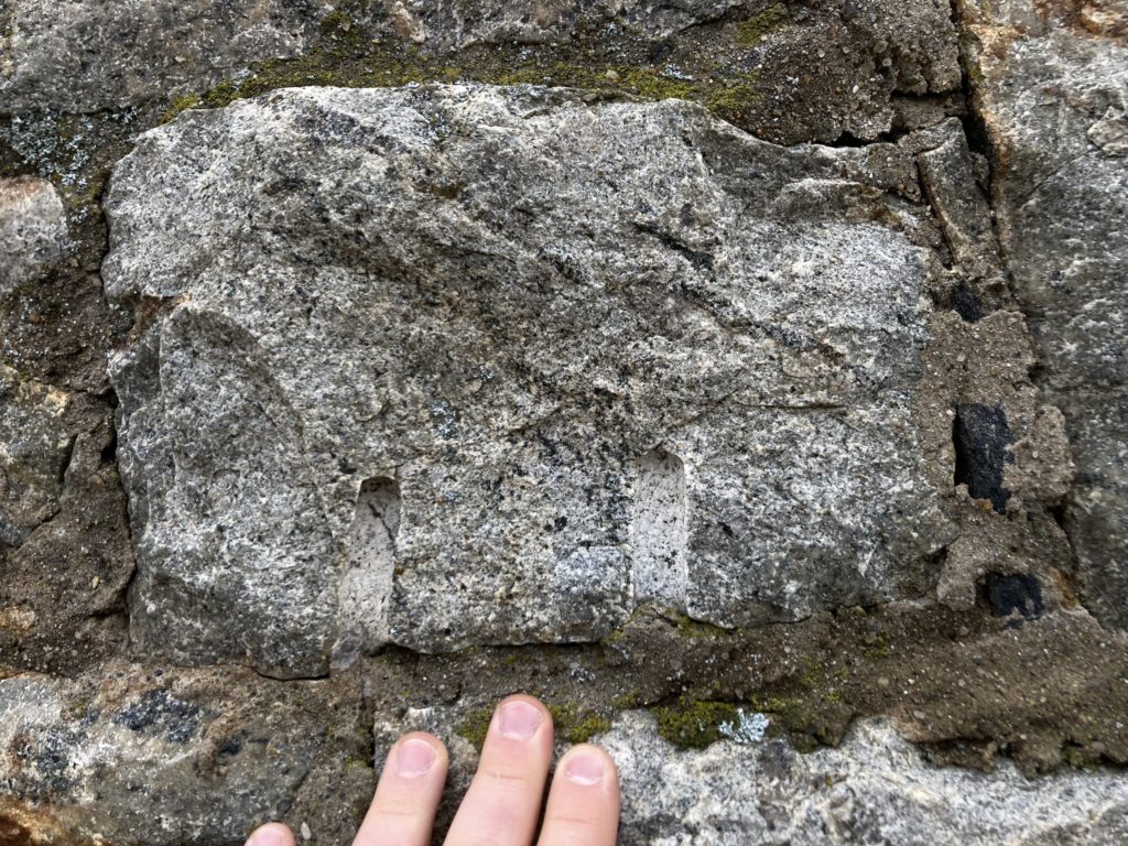 a block of rough hewn stone in the wall of a fire tower, hand for scale.  The block has 2 inch deep, half inch wide drill holes in it, which are evidence of being quarried.