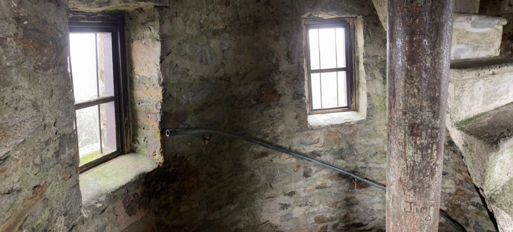 The inside of a stone tower, showing light pouring in through small windows set into the wall, with cast iron cross pieces within the windows.  A portion of the cement spiral staircase within the tower occupies the right portion of the photo