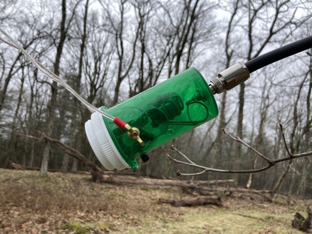 A large emerald green transparent plastic pill bottle from a pharmacy, converted into housing for a 1:1 RF choke balun. Inside the pill bottle one can see two wound toroids, wired in series to a BNC bulkhead connector from the bottom of the bottle, to which a length of RG-8U coax is connected. About 3/4 way towards the top of the bottle is a brass wing nut, which clamps down a ring terminal attached to 18AWG speaker wire, comprising one conductor of the open feedline of the antenna