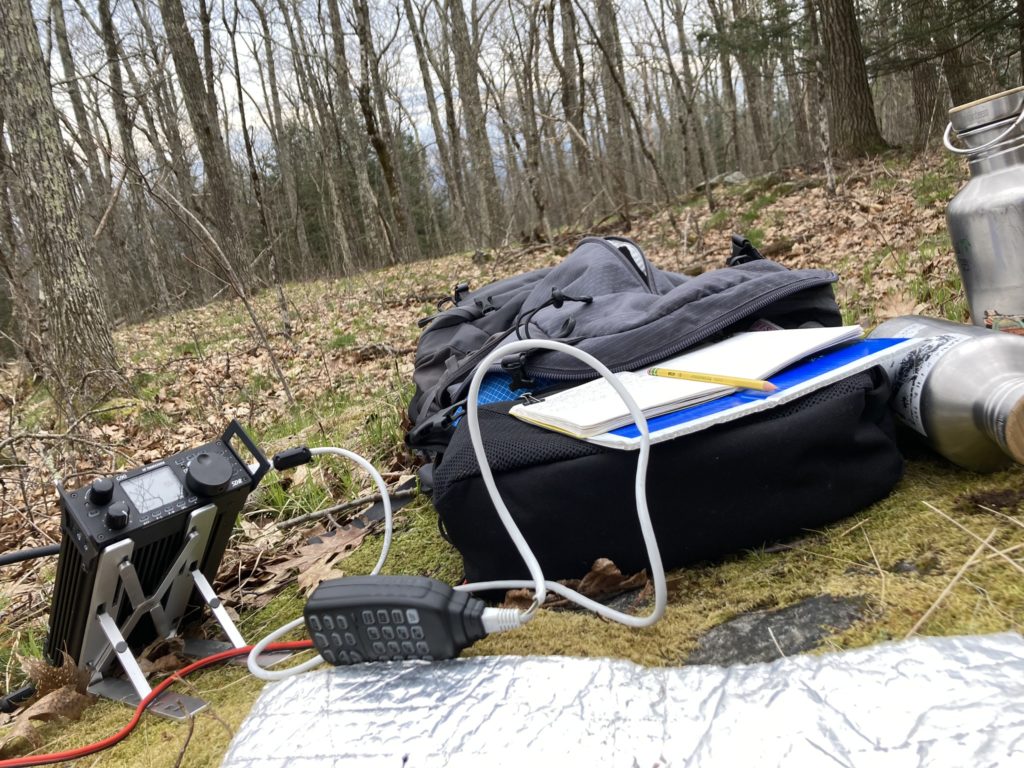 A Xiegu G90 HF transceiver, backpack, field notebook, and various miscellany lying on the ground in a bare pre-leafout New England wood.