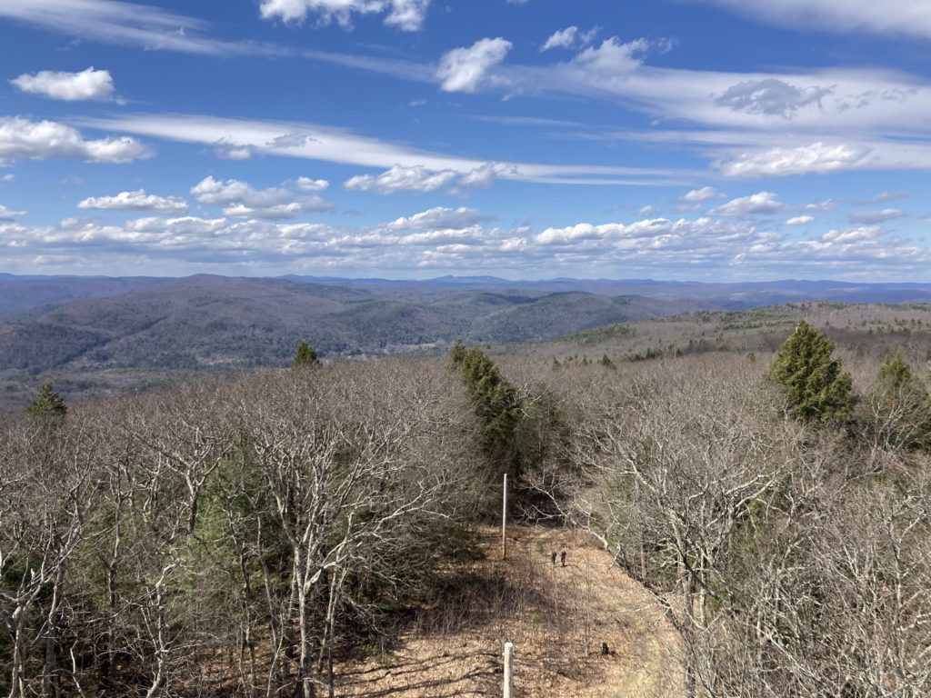 Looking down from the top of the tower towards cleared powerlines and a woods road leading down from the summit.  A landcape of New England rugged rolling hills extends into the distance under a blue sky with bright white streaky clouds.